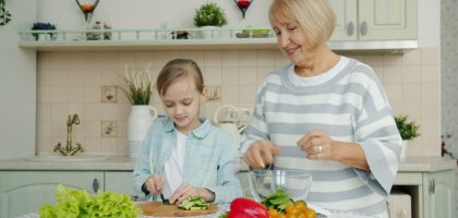 Granddaughter and grandmother are making salad together and talking in kitchen at home, little girl is cutting vegetables while old woman is watching her.