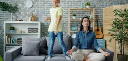 Happy kid boy is dancing on sofa while young mother is meditating in lotus position at home sitting on couch with closed eyes. Childhood and parenthood concept.