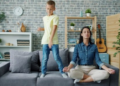 Happy kid boy is dancing on sofa while young mother is meditating in lotus position at home sitting on couch with closed eyes. Childhood and parenthood concept.